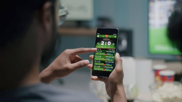 Person using a mobile sports betting app while watching a football match Person using a mobile sports betting app while watching a football match